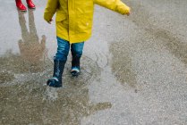 Cropped unrecognizable adorable joyful children in red and yellow raincoat and rubber boots having fun playing in puddle in street in park in gray day — Stock Photo
