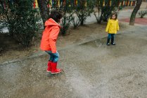 Adorables niños alegres en impermeable rojo y amarillo y botas de goma que se divierten jugando en charco en la calle en el parque en día gris - foto de stock
