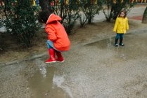 Adorable joyful children in red and yellow raincoat and rubber boots having fun playing in puddle in street in park in gray day — Stock Photo