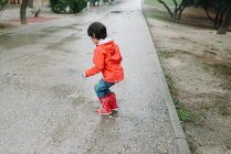 Back view of anonymous adorable joyful child in red raincoat and rubber boots having fun playing with puddle on street in park in gray day — Stock Photo