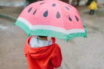 Back view of unrecognizable kid with watermelon styles open umbrella in red raincoat and rubber boots walking in park alley in gray day — Stock Photo