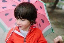 Active kid with watermelon styles open umbrella in red raincoat and rubber boots looking away in park alley in gray day — Stock Photo