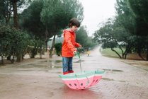 Aktives Kind mit Wassermelone im Stil öffnet Regenschirm in rotem Regenmantel und Gummistiefeln bei grauem Tag in Parkallee — Stockfoto