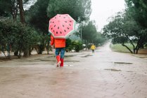 Visão traseira da criança irreconhecível com estilos de melancia guarda-chuva aberto em capa de chuva vermelha e botas de borracha andando no beco do parque no dia cinza — Fotografia de Stock