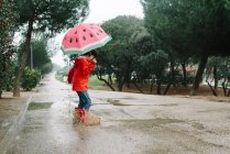 Vue latérale enfant actif avec des styles pastèque parapluie ouvert en imperméable rouge et bottes en caoutchouc sautant jouer dans l'allée du parc dans la journée grise — Photo de stock