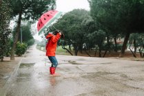 Side view active kid with watermelon styles open umbrella in red raincoat and rubber boots jumping playing in park alley in gray day — Stock Photo