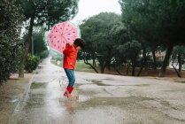 Side view active kid with watermelon styles open umbrella in red raincoat and rubber boots jumping playing in park alley in gray day — Stock Photo