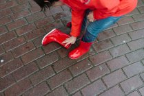 Курортна дитина поливає дощову воду з червоного гумового чоботи у вологому парку біля дерев'яної лави в сірий день — стокове фото