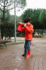 Curios kid pouring rain water from red rubber boot in wet park near wooden bench in gray day — Stock Photo