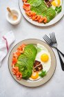 Вид зверху на тарілки з апетитом смачний барвистий здоровий сніданок, включаючи смажені яйця зі свіжим нарізаним авокадо та лососем, поміщеним на стіл з білою скатертиною — стокове фото