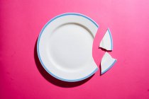 Top view of broken clean white plate with blue border and shards on gradient pink background — Stock Photo
