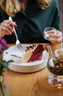 Erntehelferin in dunkelgrüner Bluse sitzt am Holztisch und isst leckeren Kuchen mit roten Beeren, serviert in weißem Keramikteller und trinkt Kräutertee — Stockfoto