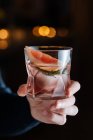 Cropped unrecognizable person hand holding glass with cold alcoholic cocktail with slice of grapefruit and ice cube placed on table against black background — Stock Photo