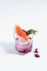 Стекло с холодным алкогольным коктейлем с ломтиком грейпфрута, розмарином, сухой черникой, цветами и кубиком льда на столе на белом фоне — стоковое фото