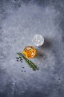 From above top view of glass cup with cold old fashioned cocktail with whiskey and orange peel placed on grey table with rosemary plant a pepper grains — Stock Photo