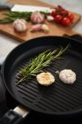 Aromatic grilled sprigs of rosemary and cut in half garlic head on grill pan placed on stove with cutting board with fresh herbs and vegetables in background — Stock Photo