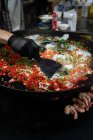 De arriba de la cosecha el cocinero en el guante el plato de la cocción del guisante con el tomate y la cebolla cerca de los langostinos en el molde metálico en el mercado - foto de stock