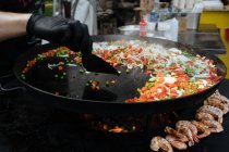 Зверху шеф-кухар в рукавичках приготування страви гороху з помідорами та цибулею біля креветок на металевій сковороді на ринку — стокове фото