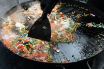 De arriba de la cosecha el cocinero en el guante el plato de la cocción del guisante con el tomate y la cebolla cerca de los langostinos en el molde metálico en el mercado - foto de stock