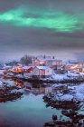 Яркие полярные огни, сияющие над симпатичной маленькой деревней на берегу отдаленной реки, окруженной белыми морозными скалами зимой в Сволваере, Норвегия — стоковое фото