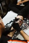 Зверху обрізаний невпізнаваний чоловік з використанням татуювальної машини, щоб зробити татуювання на нозі клієнта врожаю під час роботи в салоні — стокове фото