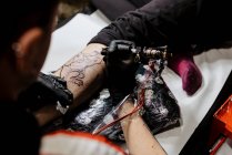From above cropped unrecognizable man with using tattoo machine to make tattoo on leg of crop customer during work in salon — Stock Photo