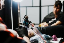 De cima cortado homem irreconhecível com o uso de máquina de tatuagem para fazer tatuagem na perna do cliente da colheita durante o trabalho no salão — Fotografia de Stock