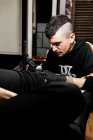 Homem elegante com piercing usando máquina de tatuagem para fazer tatuagem na perna do cliente durante o trabalho no salão — Fotografia de Stock