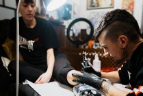 Hombre con estilo con piercing utilizando la máquina de tatuaje para hacer tatuaje en la pierna del cliente de la cosecha durante el trabajo en el salón - foto de stock