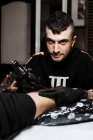 Стильный счастливый мужчина с блестками смотрит в камеру при помощи тату-машины, чтобы сделать татуировку на ноге анонимного клиента во время работы в салоне — стоковое фото