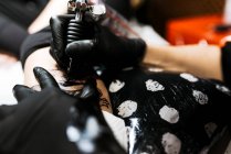 De arriba recortado hombre irreconocible con el uso de la máquina de tatuaje para hacer tatuaje en la pierna del cliente de la cosecha durante el trabajo en el salón - foto de stock