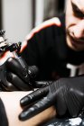 Hombre con estilo con piercing utilizando la máquina de tatuaje para hacer tatuaje en la pierna del cliente de la cosecha durante el trabajo en el salón - foto de stock