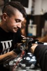 Homem elegante com piercing usando máquina de tatuagem para fazer tatuagem na perna do cliente durante o trabalho no salão — Fotografia de Stock