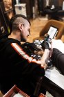 De cima homem elegante com piercing usando máquina de tatuagem para fazer tatuagem na perna do cliente de colheita durante o trabalho no salão — Fotografia de Stock