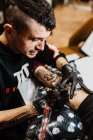 Сверху стильный мужчина с пирсингом с помощью тату-машины, чтобы сделать татуировку на ноге клиента урожая во время работы в салоне — стоковое фото