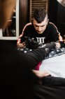 Homme élégant avec perçage à l'aide d'une machine à tatouer pour faire du tatouage sur la jambe du client de la culture pendant le travail dans le salon — Photo de stock