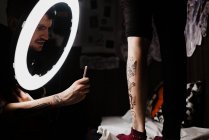 Неузнаваемый мужчина-татуировщик, использующий смартфон, чтобы сфотографировать татуировку на ноге клиента для портфолио в современной студии — стоковое фото