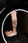 Crop irriconoscibile gamba donna con tatuaggio fresco in lampada rotonda incandescente durante la sessione fotografica nel salone di tatuaggio — Foto stock