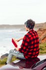 Back view of faceless extravagant lady in casual wear and bandana sitting on hood of car and reading book at empty seaside — Stock Photo