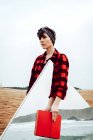 Jeune femme sérieuse et coûteuse en chemise à carreaux rouge et noire décontractée debout sur une plage de sable avec livre rouge à la main et grand miroir avec réflexion sur la mer et les rochers — Photo de stock