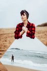 Thoughtful sad young female in casual checkered shirt standing on sandy beach and holding large mirror with reflection of stormy sea and walking man — Stock Photo