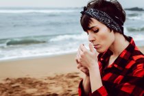 Вид сбоку на молодую неформальную женщину в клетчатой рубашке и повязке на голове, зажигая сигарету, стоя на песчаном пляже с бурным морем на заднем плане — стоковое фото