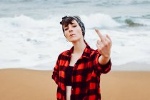Mulher jovem desafiante arrogante com piercing e cigarro vestindo camisa desabotoada mostrando o dedo médio enquanto estava em pé na praia de areia com ondas do mar no fundo — Fotografia de Stock