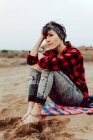 Tranquillo rilassato pensieroso hipster femminile in jeans strappati e camicia a scacchi seduto sulla spiaggia sabbiosa in giornata nuvolosa — Foto stock