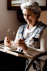 Розслаблена зріла жінка з сірим волоссям і доброю посмішкою читає книгу тримає окуляри в руці біля вікна в кріслі — стокове фото