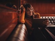 Стильна розумна щаслива жінка, серфінг планшет, зручно сидить з ногами на чорному шкіряному дивані в кафе — стокове фото