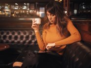 Elegante inteligente mujer reflexiva surf tableta cómodamente sentado en el sofá de cuero negro en la cafetería con la tableta y beber café en una taza - foto de stock