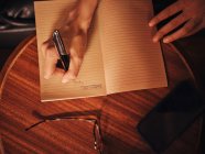 Зверху врожаю невизначена жінка пише з ручкою в блокноті за круглим дерев'яним столом з мобільним телефоном і окулярами в кафе — стокове фото