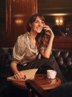 Mujer feliz con estilo escuchando atentamente la información en el teléfono móvil y la escritura en el portátil cómodamente sentado en la mesa redonda en la cafetería - foto de stock