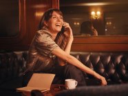 Стильная счастливая женщина внимательно слушает информацию по мобильному телефону и пишет в блокноте комфортно сидя за круглым столом в кафе — стоковое фото